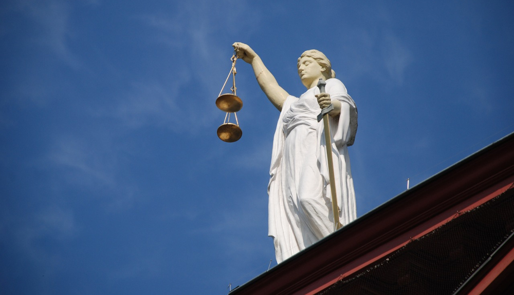 A statue of lady justice is seen in front of  a blue sky. she is blindfolded. in one hand she holds scales, and in the other, a sword.