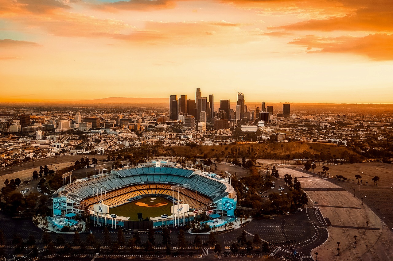 overhead view of Los Angeles at sunset, the cityscape lit up orange beneath an orange sky. in the forefront is LA Dodgers Stadium. 