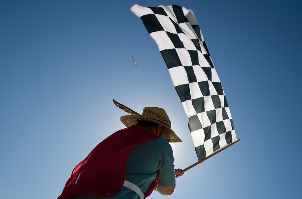 person waves a checkerboard flag in the air, seen from behind and below. they wear a red cape and a hat. they are illuminated by the sun in front of them. 
