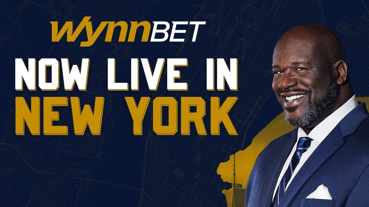 Shaquille O'neal, an attractive bald black basketball player in a blue suit, smiles in front of a blue background with text that reads "WynnBet Now Live in New York" to advertise the sportsbook's launch in NY state this week.