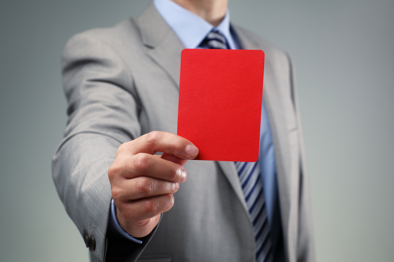 a man in a grey suit is seen from the neck down. he holds a red penalty card towards the camera.