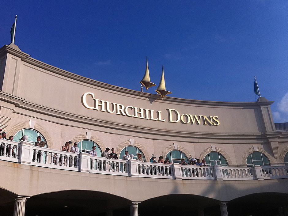 The balcony at Churchill Downs racetrack is seen against a blue sky as spectators stand and watch. Historic Churchill Downs Inc just acquired even more racing properties, including Colonial Downs, VA's only racetrack.