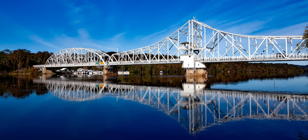 The East Haddam Bridge in Haddam Connecticut is seen stretching over sparkling water, its reflection shimmering back in the river below. Beautiful blue skies above.
