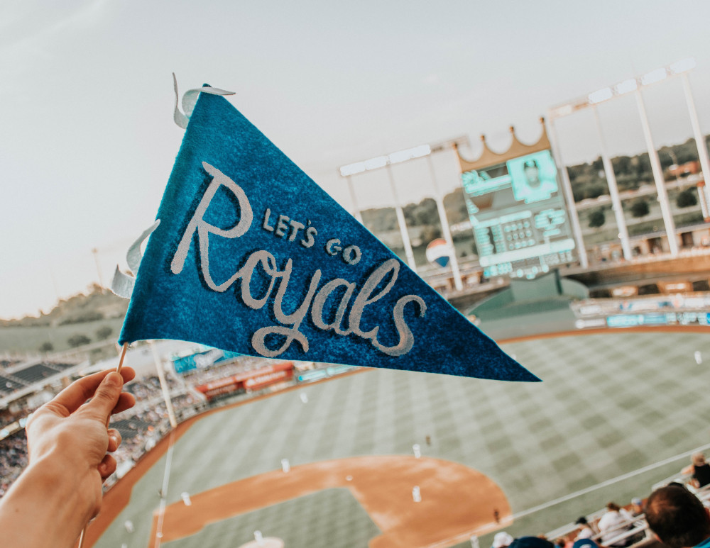 a hand holds up a "let's go Royals!" pennant at a Kansas City Royals baseball game. The field is seen in the background. Kansas sports betting bill would earmark 80% of tax revenue for incentives to lure pro sports teams to KS..