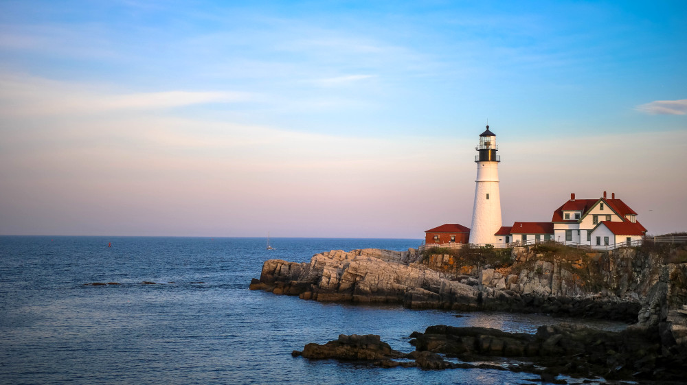 Portland Head Light lighthouse in Maine is seen at sunrise, the sky a beautiful blue fading into yellow, pink, and orange as the lighthouse sits atop a cliff with the water below.