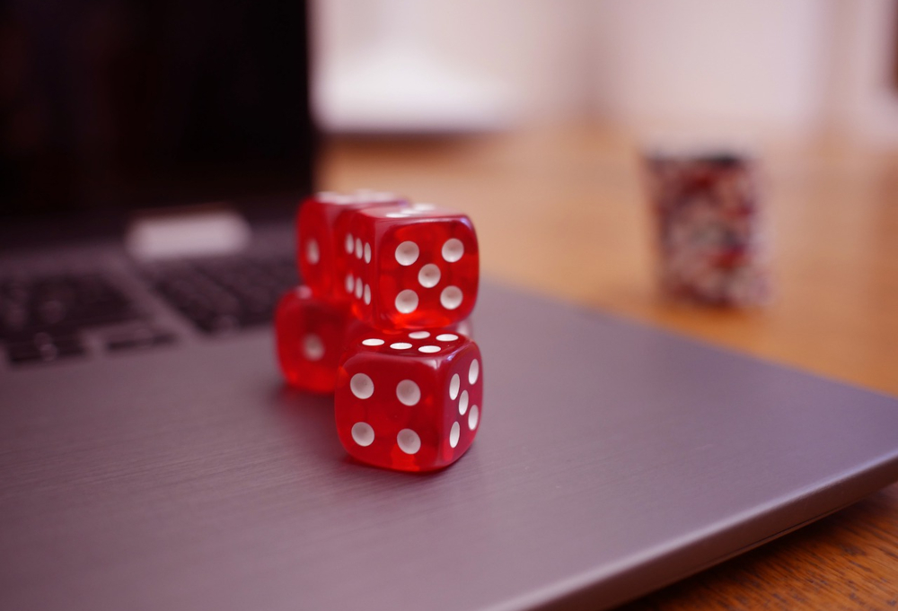 A closeup of a laptop with 4 red die near the edge, under the keyboard. in the background behind it is a stack of casino/poker chips.