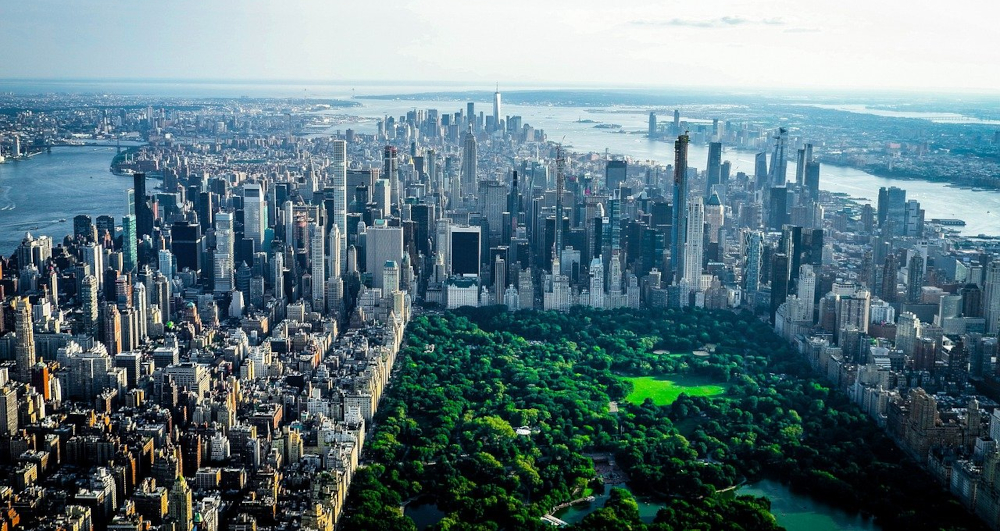 Overhead view of Manhattan in NYC -- a rectangle of green trees in Central Park and then rows and rows of skyscrapers and buildings of various heights. On the horizon, water separating lower manhattan from new jersey. 