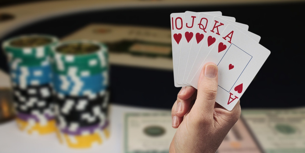 Photo of poker player's hand, holding cards 10JQKA of hearts. Blurry poker chips and cash on the poker table past his hand. Throughout the past 2 years of COVID-19, legal online poker has heated up as in-person games became scarce.
