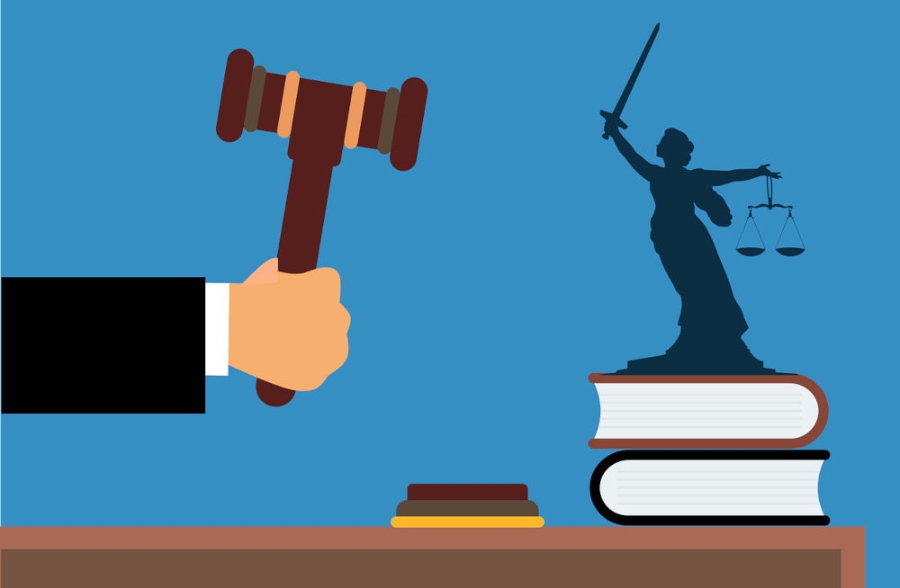 illustration of a judge's hand holding a gavel. on the table is also a statue of blind justice holding her scales and a stack of books. we await the judge's decision on the DOJ's motion to dismiss its IGT lawsuit.